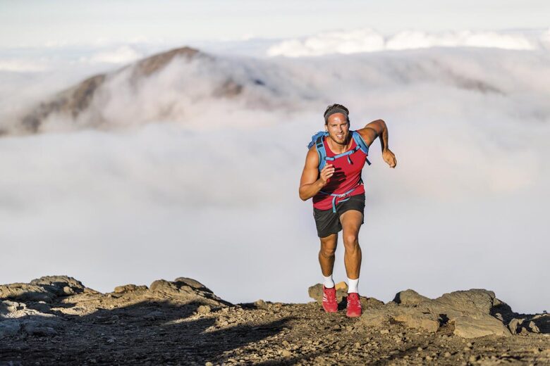 Runner trail running fitness man on endurance run motivation and concentration on race in sky and clouds background on nature landscape. Focused athlete with backpack and hairband training cardio.