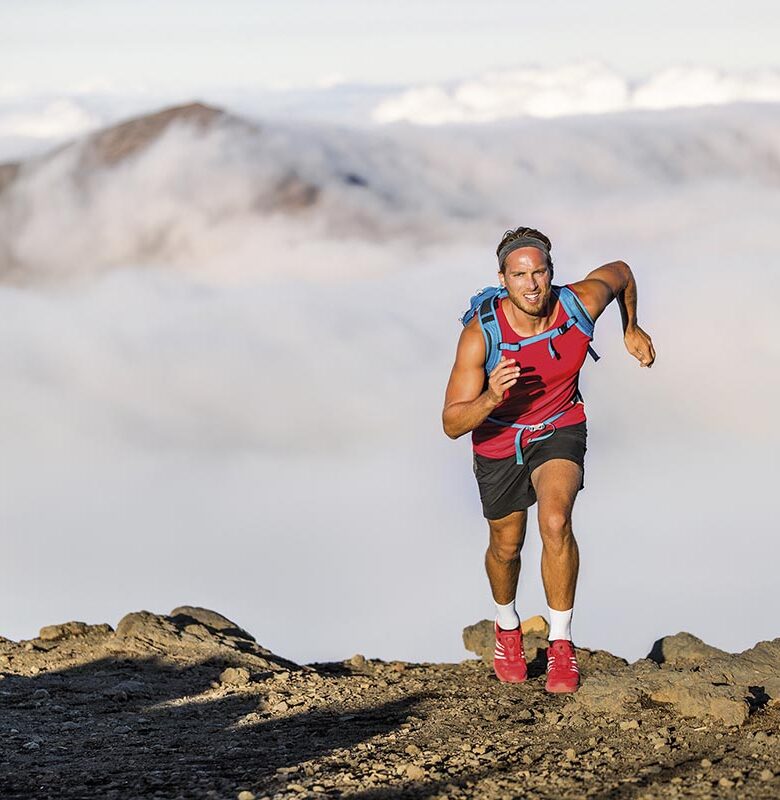 Runner trail running fitness man on endurance run motivation and concentration on race in sky and clouds background on nature landscape. Focused athlete with backpack and hairband training cardio.