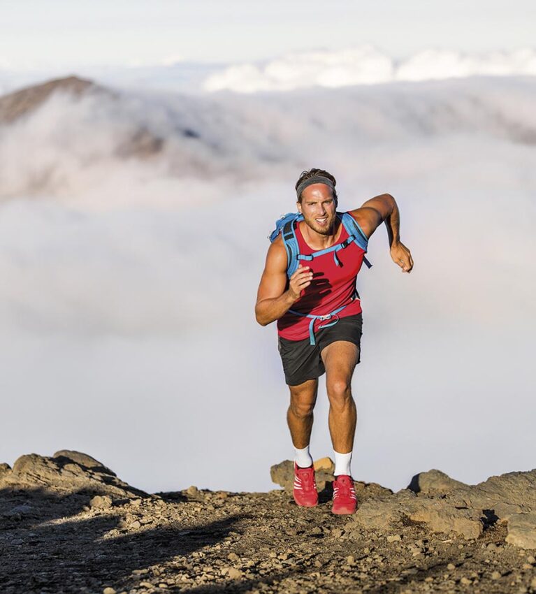 Runner trail running fitness man on endurance run motivation and concentration on race in sky and clouds background on nature landscape. Focused athlete with backpack and hairband training cardio.