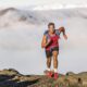 Runner trail running fitness man on endurance run motivation and concentration on race in sky and clouds background on nature landscape. Focused athlete with backpack and hairband training cardio.