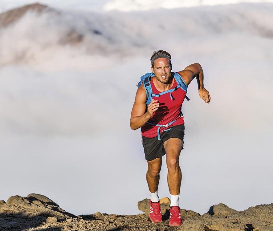 Runner trail running fitness man on endurance run motivation and concentration on race in sky and clouds background on nature landscape. Focused athlete with backpack and hairband training cardio.